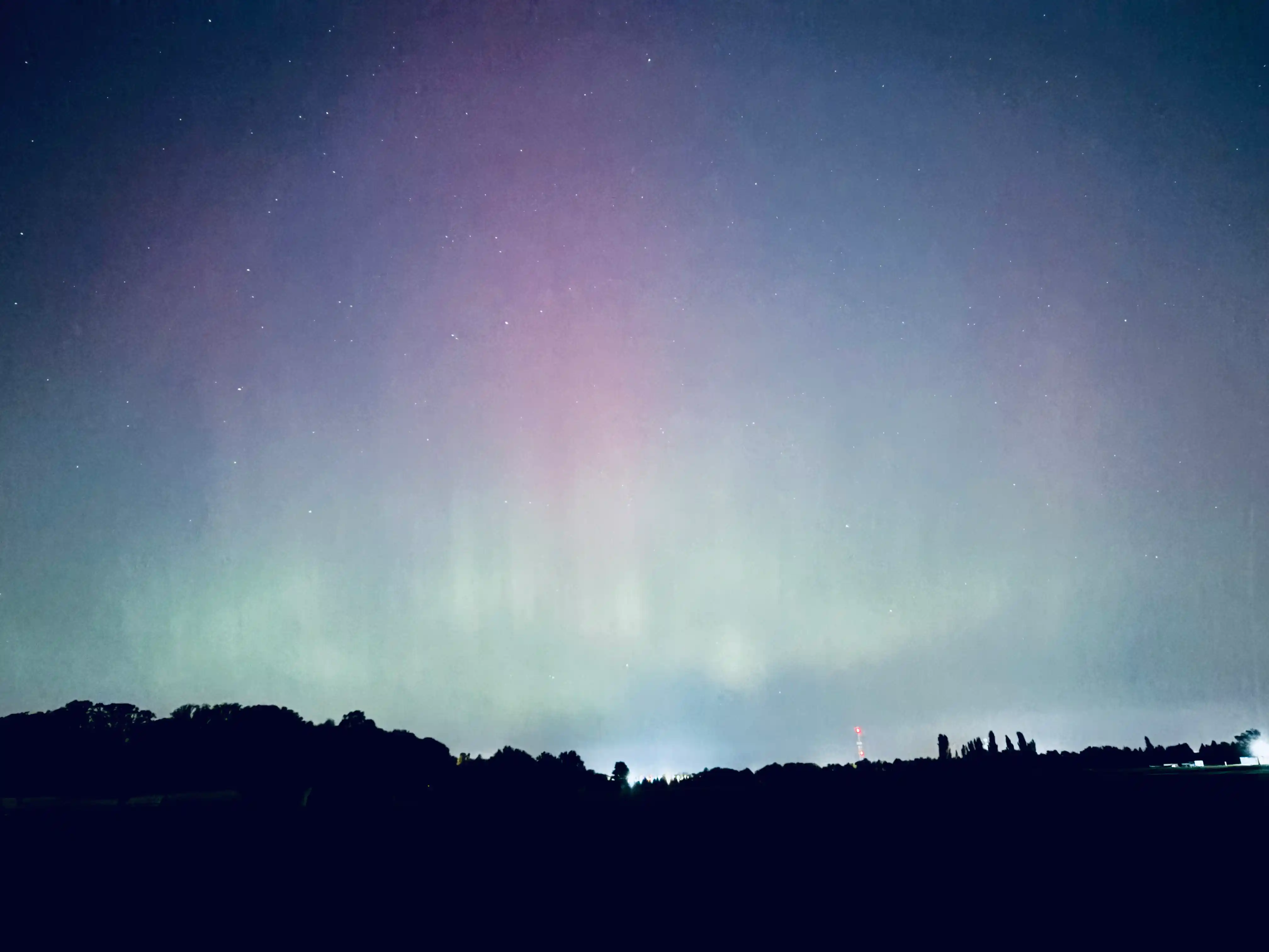 This photo captures the mesmerizing phenomenon of the aurora borealis illuminating the night sky above a dark, silhouetted landscape. The sky is filled with soft, glowing bands of light, predominantly in ethereal shades of pale green and light purple, which appear to shimmer and spread across the upper portion of the frame. Numerous small, faint stars are visible through the auroral glow, adding to the celestial beauty seen from Lafayette, Indiana.

Along the horizon, a dark, irregular profile of a distant treeline forms the lower boundary of the image, with variations in height. This dark foreground emphasizes the vibrancy of the sky. In the far distance, subtle glows of artificial light, possibly from a distant part of Lafayette or a neighboring area, can be discerned, along with the faint outline of what appears to be a communication tower or spire. The overall impression is one of profound natural wonder and the quiet majesty of a night sky transformed by an atmospheric light show in the Indiana sky.