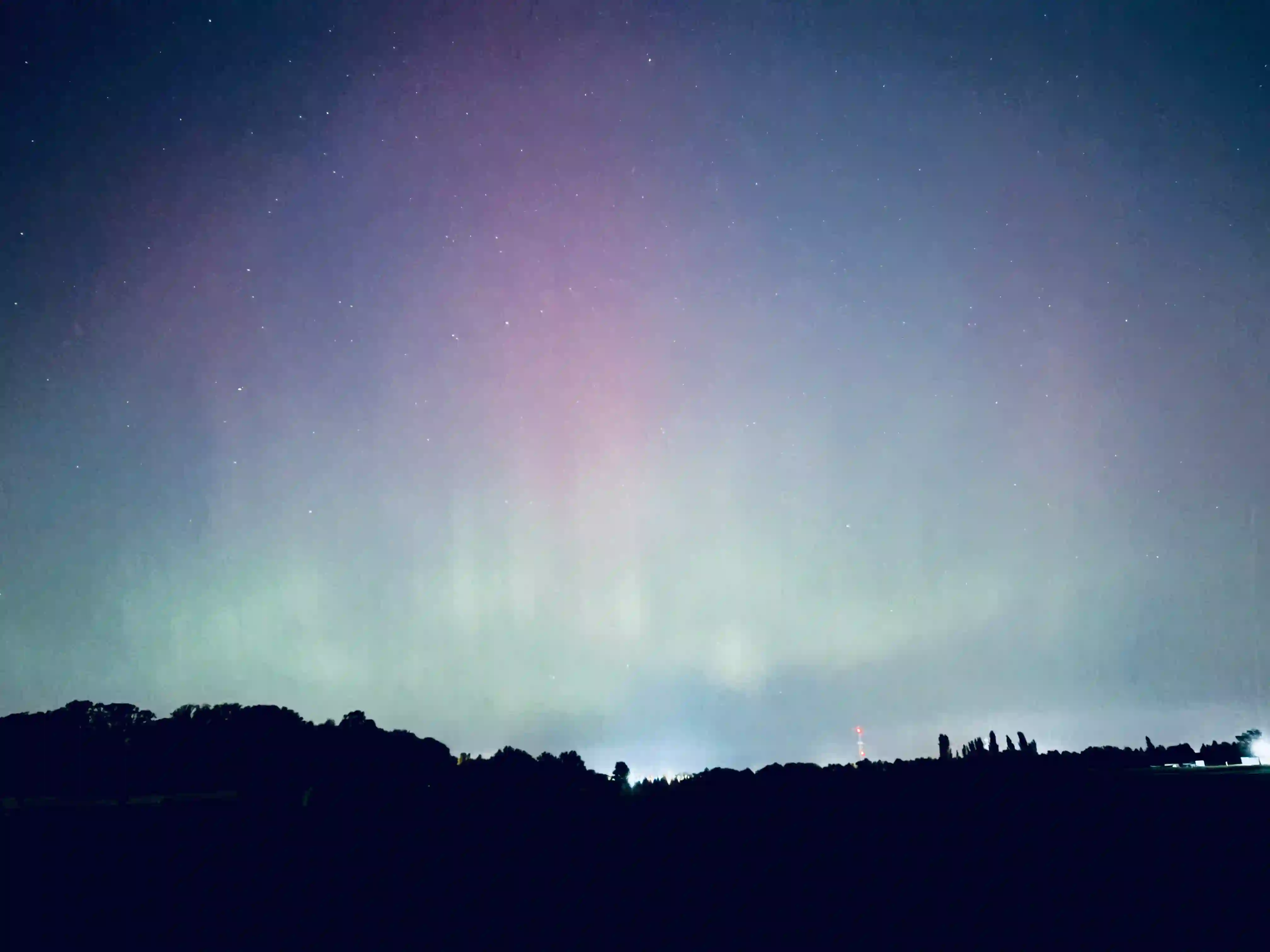 This photo captures the mesmerizing phenomenon of the aurora borealis illuminating the night sky above a dark, silhouetted landscape. The sky is filled with soft, glowing bands of light, predominantly in ethereal shades of pale green and light purple, which appear to shimmer and spread across the upper portion of the frame. Numerous small, faint stars are visible through the auroral glow, adding to the celestial beauty seen from Lafayette, Indiana.

Along the horizon, a dark, irregular profile of a distant treeline forms the lower boundary of the image, with variations in height. This dark foreground emphasizes the vibrancy of the sky. In the far distance, subtle glows of artificial light, possibly from a distant part of Lafayette or a neighboring area, can be discerned, along with the faint outline of what appears to be a communication tower or spire. The overall impression is one of profound natural wonder and the quiet majesty of a night sky transformed by an atmospheric light show in the Indiana sky.