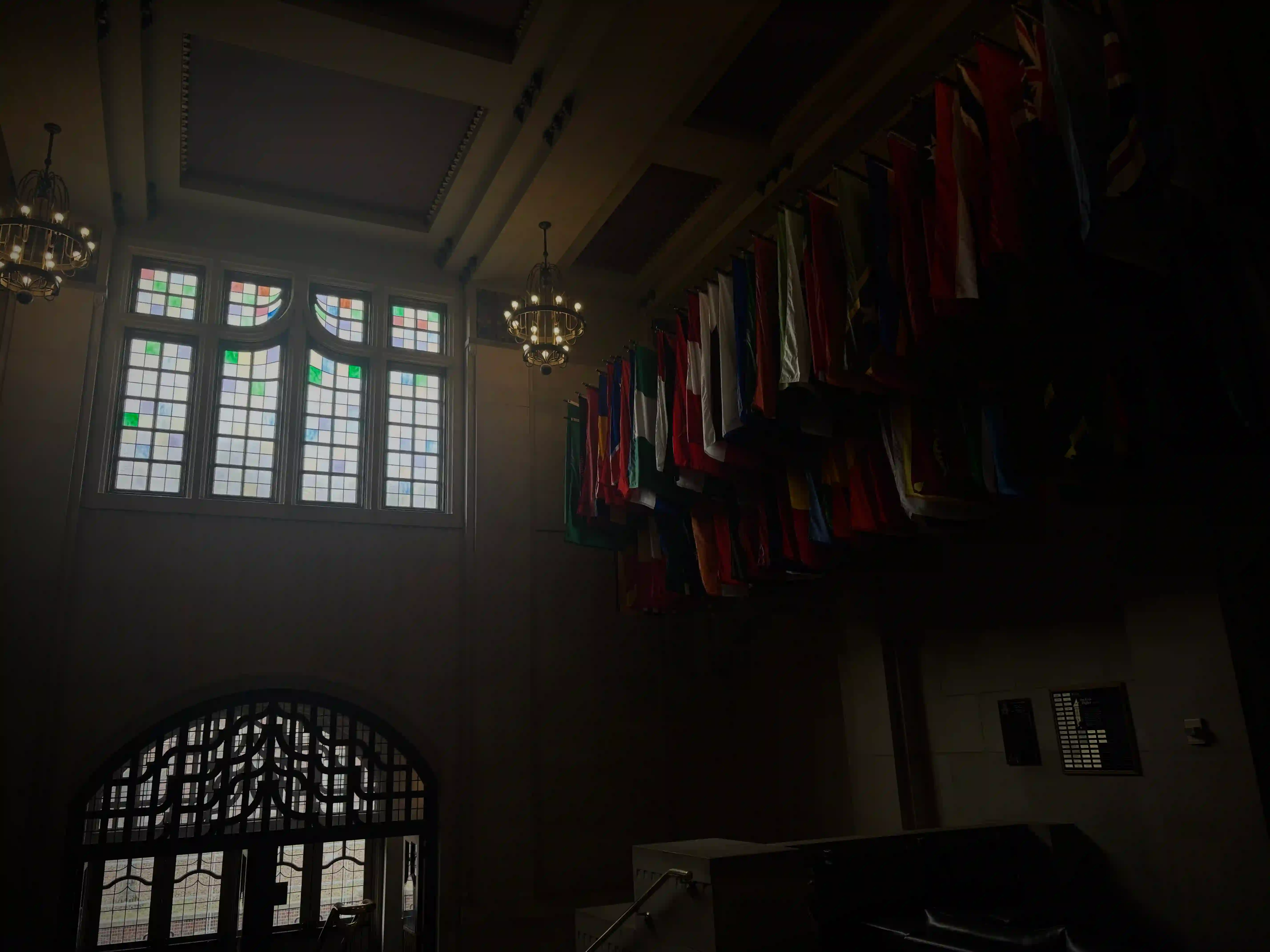 This photo captures an interior view of a grand hall within the Purdue Memorial Union, characterized by its lofty ceilings, decorative elements, and a display of international flags. In the upper right portion of the image, a long row of national flags from various countries hangs prominently from a high beam, their vibrant colors – reds, greens, blues, whites, and yellows – providing a striking contrast against the muted tones of the architecture. They appear to be hanging in a neat, uniform arrangement.

In the background, on the left side, large, ornate stained-glass windows are visible, through which soft sunlight filters, casting a gentle illumination into the space. The windows feature multiple panes, some with subtle color variations, and a classic, grid-like design. Below these windows, a large, dark arched doorway with intricate, possibly wrought-iron, grilles is visible, hinting at another passage or room.

The ceiling above is high and features decorative molding. Two ornate chandeliers with multiple light sources hang from the ceiling, adding to the stately ambiance, though their illumination is soft and diffused. The overall lighting in the room is somewhat dim, creating areas of deep shadow, particularly on the right side and in the foreground, which accentuates the light from the windows and the colors of the flags. The scene evokes a sense of historical grandeur and international presence within the PMU.