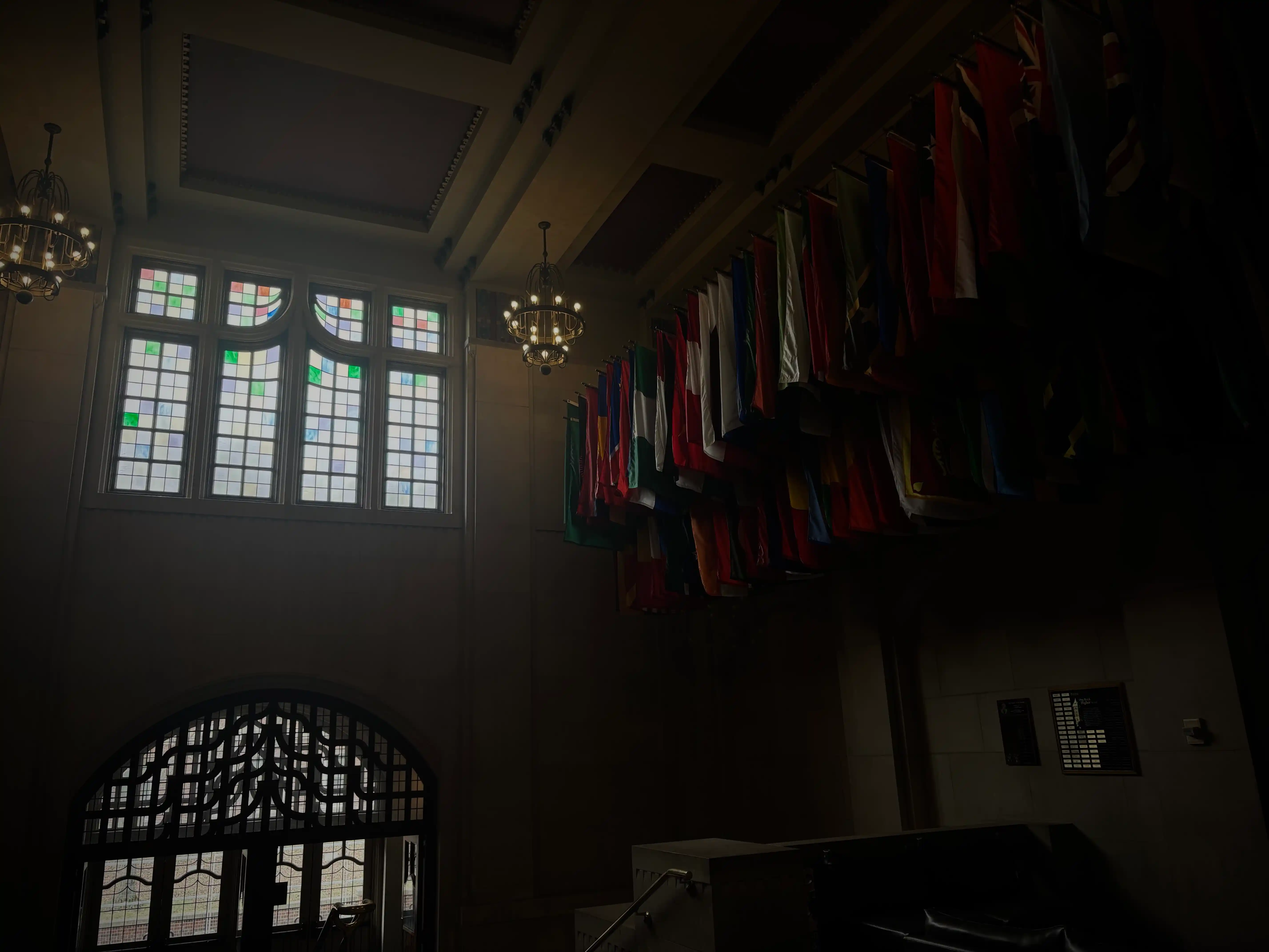This photo captures an interior view of a grand hall within the Purdue Memorial Union, characterized by its lofty ceilings, decorative elements, and a display of international flags. In the upper right portion of the image, a long row of national flags from various countries hangs prominently from a high beam, their vibrant colors – reds, greens, blues, whites, and yellows – providing a striking contrast against the muted tones of the architecture. They appear to be hanging in a neat, uniform arrangement.

In the background, on the left side, large, ornate stained-glass windows are visible, through which soft sunlight filters, casting a gentle illumination into the space. The windows feature multiple panes, some with subtle color variations, and a classic, grid-like design. Below these windows, a large, dark arched doorway with intricate, possibly wrought-iron, grilles is visible, hinting at another passage or room.

The ceiling above is high and features decorative molding. Two ornate chandeliers with multiple light sources hang from the ceiling, adding to the stately ambiance, though their illumination is soft and diffused. The overall lighting in the room is somewhat dim, creating areas of deep shadow, particularly on the right side and in the foreground, which accentuates the light from the windows and the colors of the flags. The scene evokes a sense of historical grandeur and international presence within the PMU.