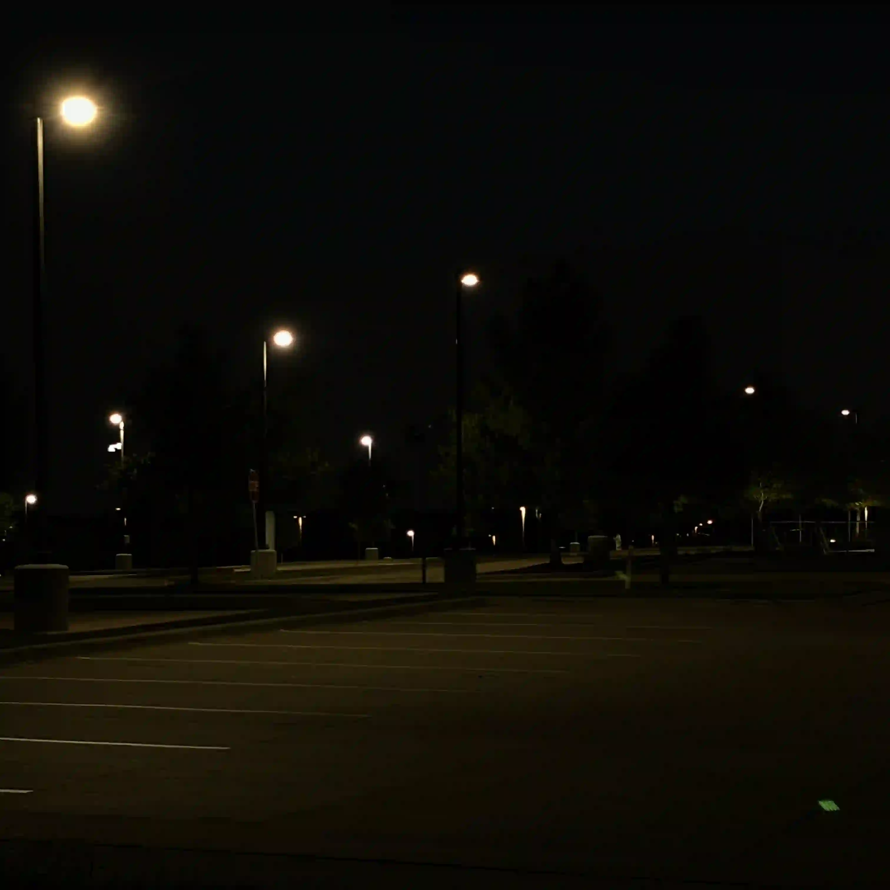This photo depicts an empty parking lot at night, illuminated by several dim streetlights, creating a somewhat eerie and desolate atmosphere. The foreground and mid-ground are dominated by the dark asphalt surface of the parking lot, marked with white painted lines for parking spaces, which appear mostly empty.

Several tall streetlights with glowing, circular luminaires cast pools of light across the lot, their beams extending into the darkness. The light sources themselves appear as bright, slightly diffused orbs. Beyond the paved area, in the background, a line of dark trees and indistinct shrubbery is visible, vaguely illuminated by the streetlights or other distant light sources, their forms mostly swallowed by the night. Some light poles are also visible further back, extending into the depth of the image. The sky above is entirely dark, adding to the pervasive sense of night. The overall mood is one of stillness and quiet, emphasized by the absence of cars and people.