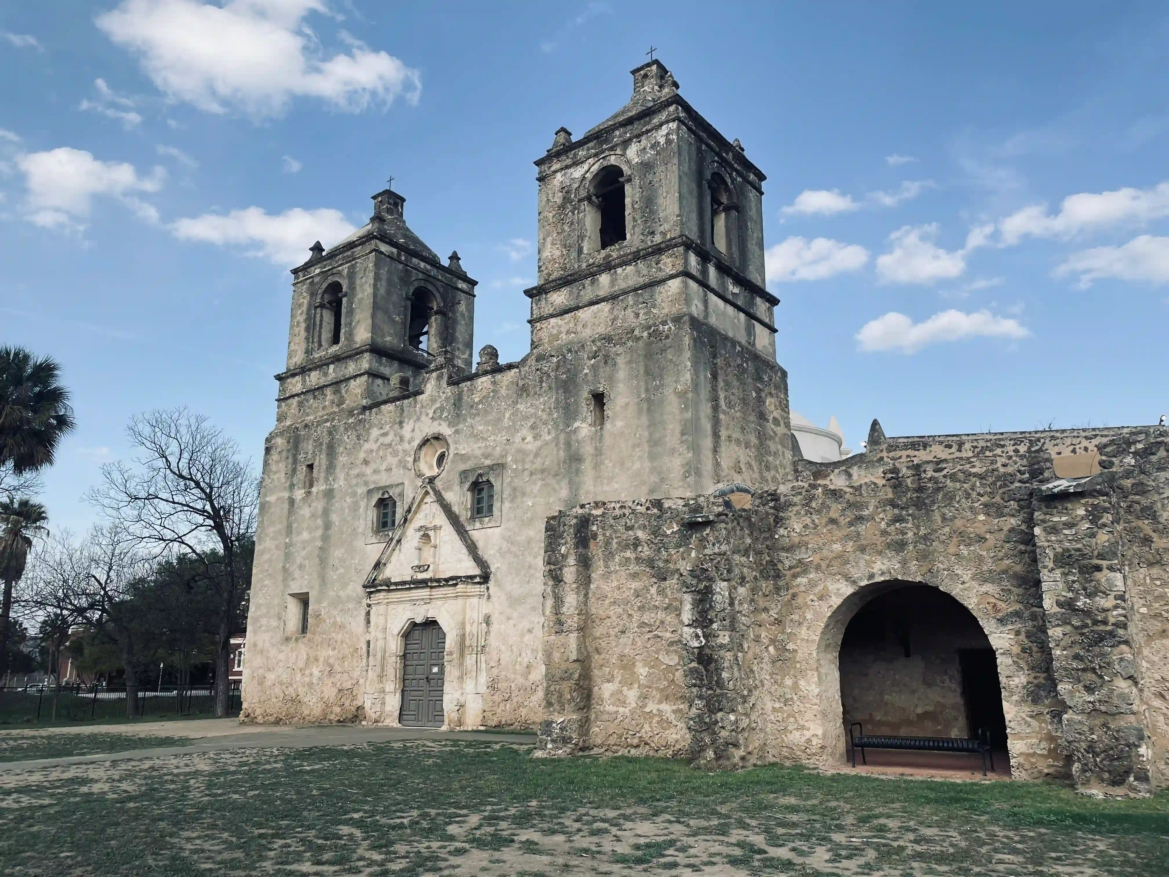 This photo showcases the Church at Mission Nuestra Señora de la Purísima Concepción de Acuña, a historic stone building under a partly cloudy sky. In the foreground, the ground is covered with sparse patches of green grass and dirt, indicating a well-trodden area. Behind this foreground, the substantial, aged stone structure of the church rises. Its weathered facade, a mottled grey-brown, displays the passage of time. The building is characterized by two prominent bell towers situated towards the center of the image, each with arched openings for bells and topped with small crosses. Below the towers, a double-doored entrance, set within an ornate, carved stone frame, leads into the church. To the right side of the building, a large stone archway is visible, suggesting an open passage or alcove within the structure, beneath which a dark bench is placed. On the far left, a tall palm tree and the bare branches of a deciduous tree are visible against the sky, adding to the sense of an established, historical site. The overall impression is one of enduring history and architectural strength.
